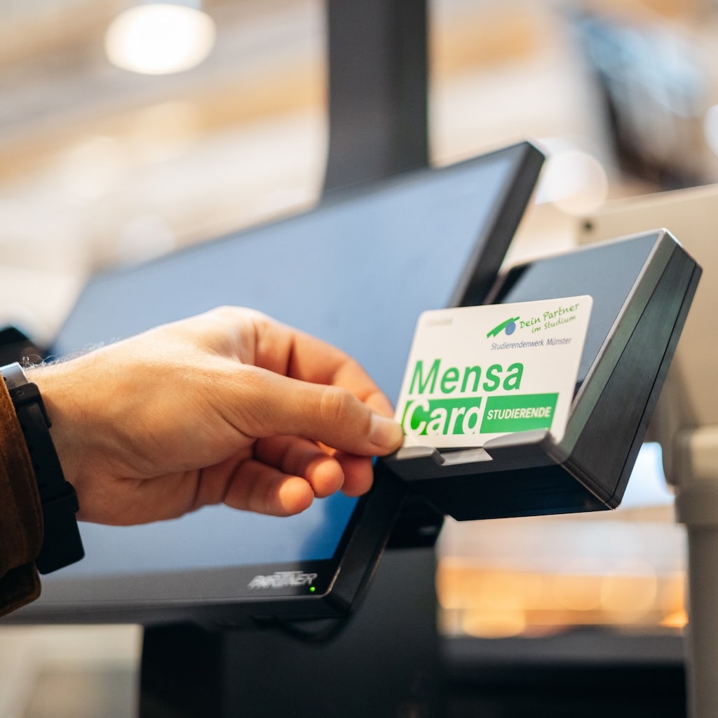 Eine Person hält eine Mensa-Karte an ein Kartenlesegerät in der Nähe eines Computerbildschirms, wahrscheinlich um eine Zahlung vorzunehmen oder eine Dienstleistung in einer Mensa oder Einrichtung in Anspruch zu nehmen.