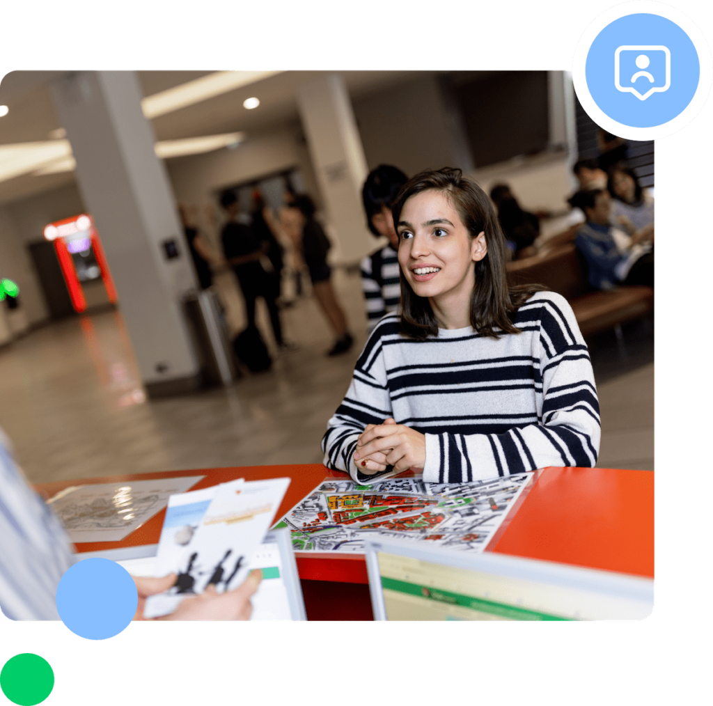 A young woman in a striped sweater stands at the info point counter, smiling and talking to someone holding brochures. There are colorful papers on the desk and people can be seen in the background of a brightly lit interior.