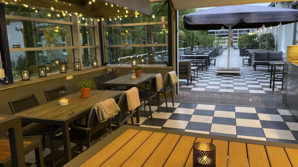 Deck 11's outdoor area with checkered floor, wooden tables, black chairs, candles and fairy lights. Potted plants adorn the tables and a covered terrace leads to further outdoor seating. Evening ambience.