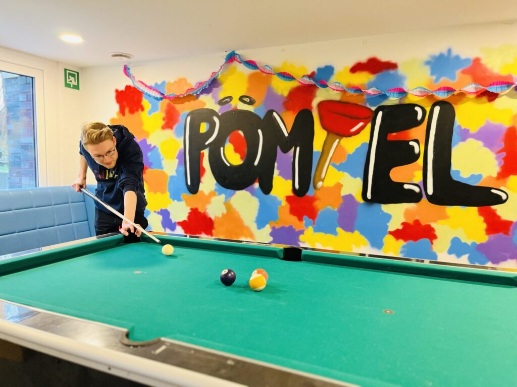 A person playing billiards aims their cue at the cue ball on a green pool table. The colorful wall behind them shows graffiti art with the word PÖMPEL and colorful decorations.