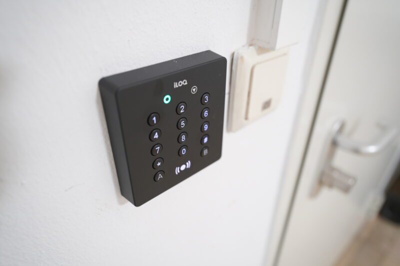 A black electronic keypad with illuminated digits is mounted on a white wall next to a door handle and a rectangular switch. There is a small indicator light above the keypad.