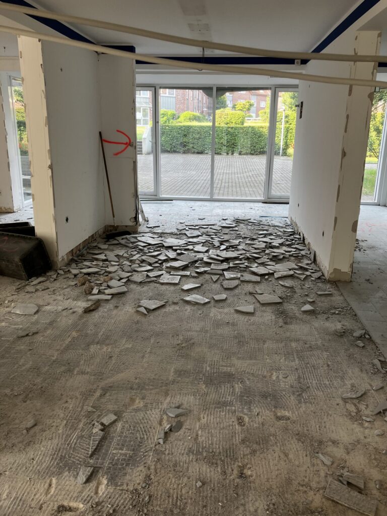 A renovated room with broken tiles on the floor, exposed walls and a red arrow on the wall pointing to the left. Large glass doors at the other end of the room lead to an inner courtyard and a green area.