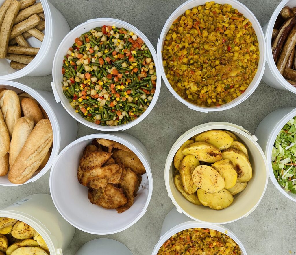 Buckets of various dishes on a table, including buns, sausages, mixed vegetables, seasoned potato slices, fried chicken pieces and a yellow rice or cereal dish with vegetables.