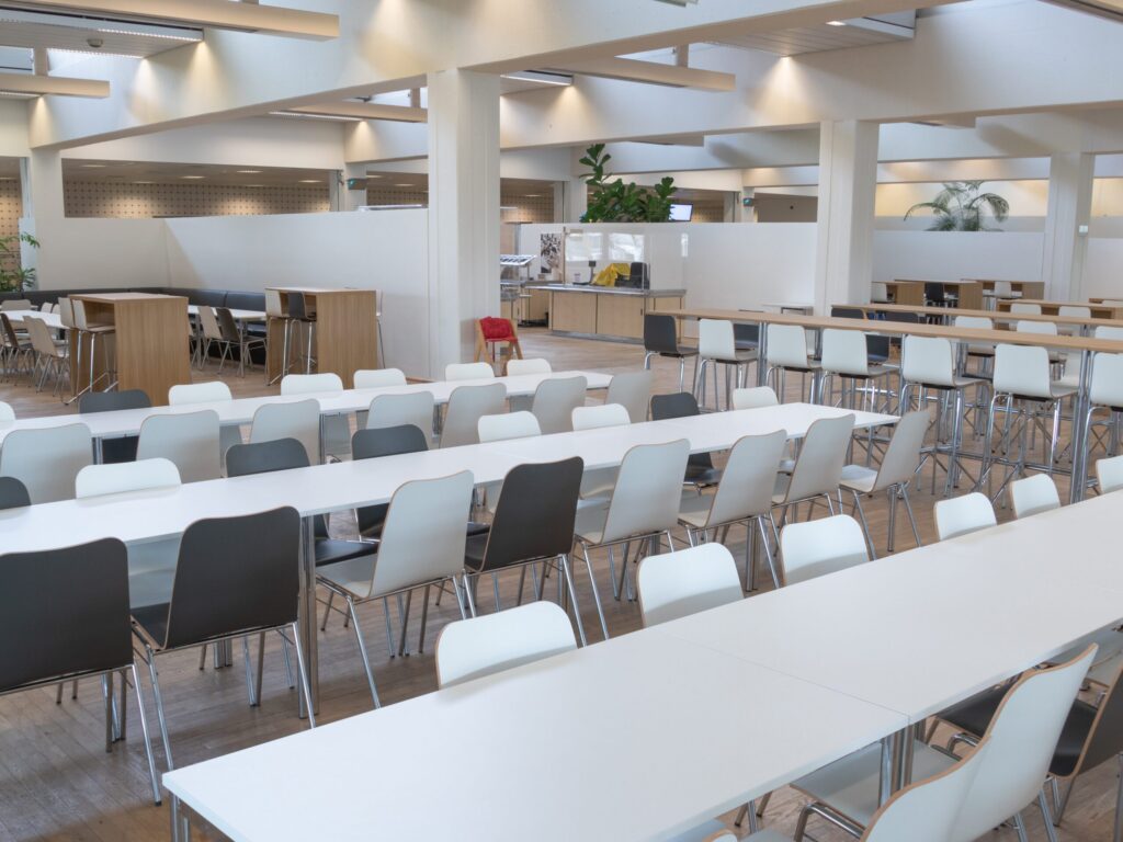 A spacious, modern cafeteria with long white tables and rows of black, gray and white chairs. The room is bright with natural light, and in the background there are plants and a food serving area.