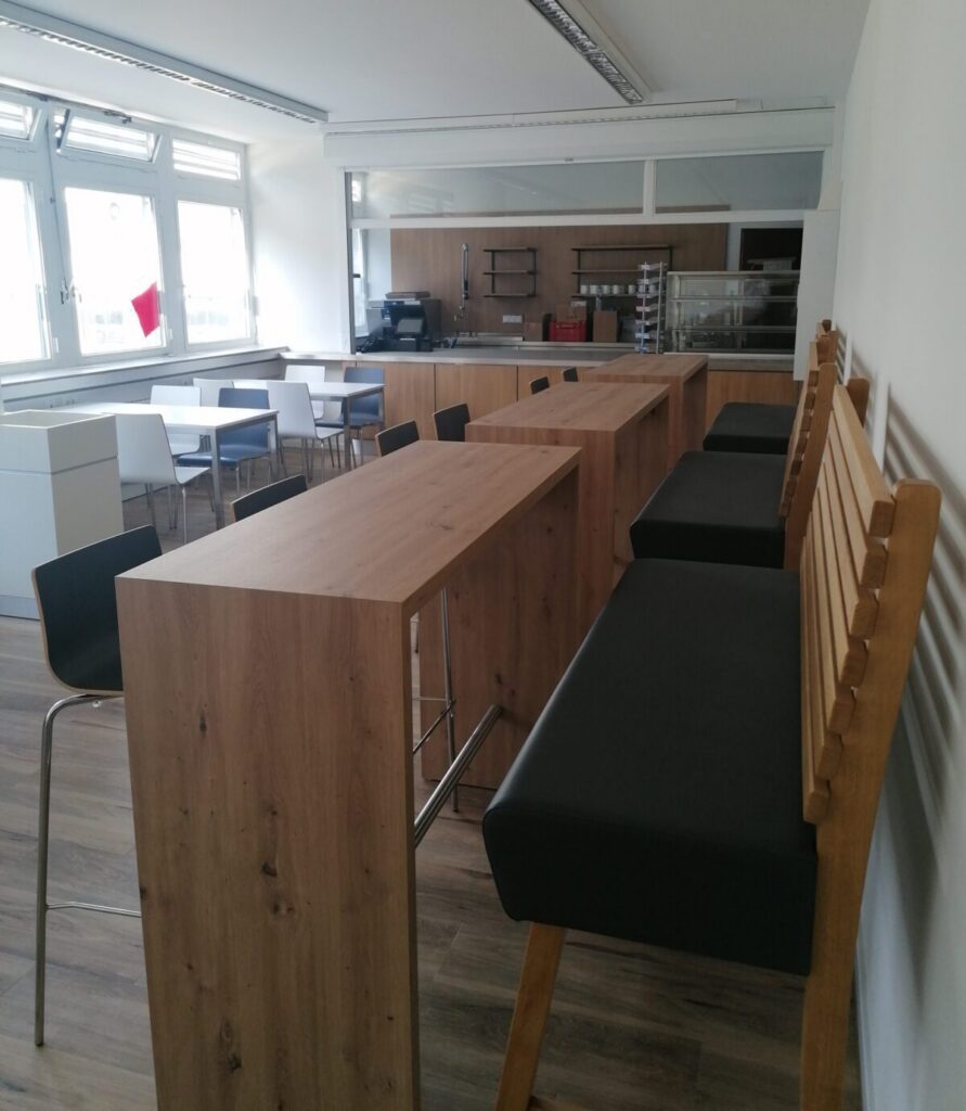 A modern, bright cafeteria with wooden bar tables, black upholstered bar stools, square tables with black chairs, large windows and a kitchen area at the back.