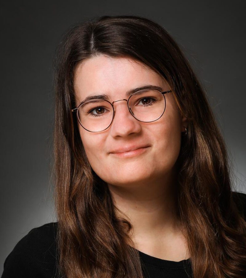 A woman with long brown hair and glasses smiles gently at the camera. She is wearing a black top and the background is a plain, dark gray gradient.