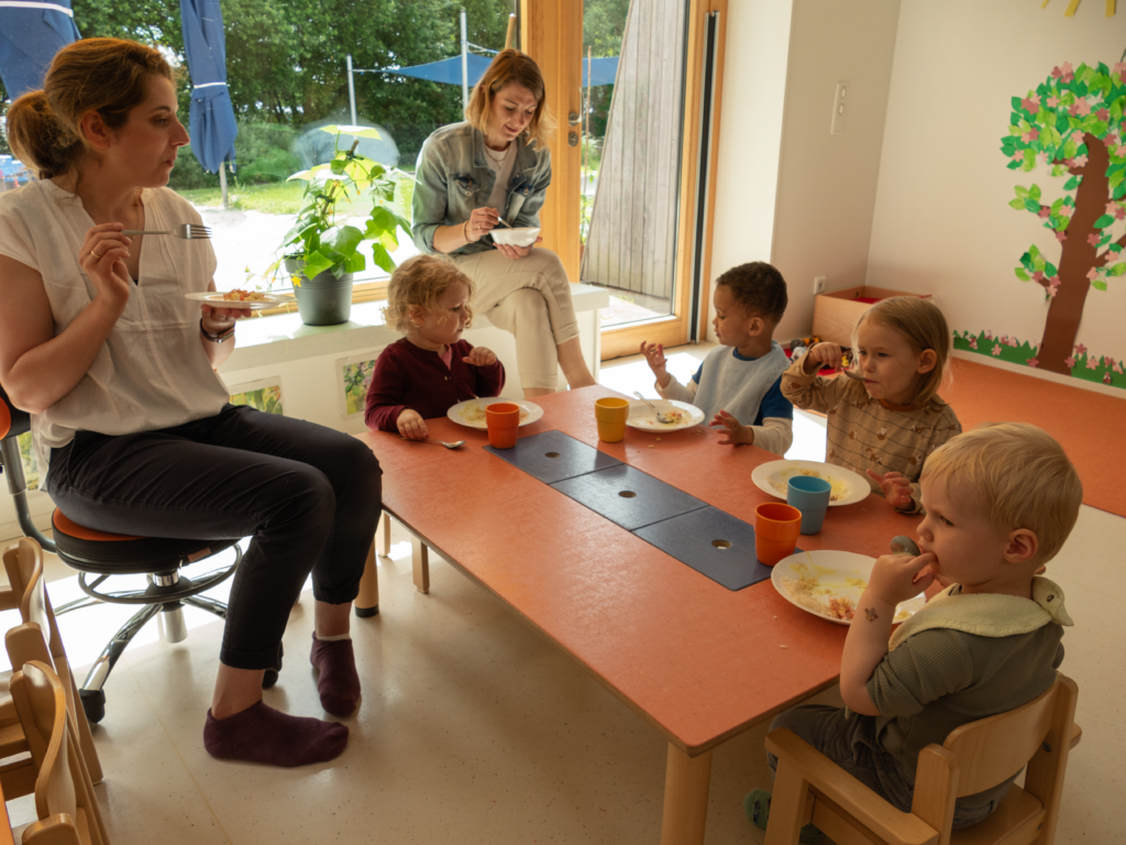 Fünf kleine Kinder sitzen um einen niedrigen Tisch und essen, während zwei Erwachsene sie beaufsichtigen. Durch die großen Fenster strömt Sonnenlicht, und der Raum ist mit Pflanzen und farbenfroher Wandmalerei dekoriert, was eine warme, fröhliche Atmosphäre schafft.