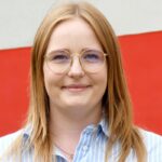 Eine Frau mit glattem, hellbraunem Haar, einer runden Brille und einem blau gestreiften Hemd lächelt vor einem rot-weißen Hintergrund.