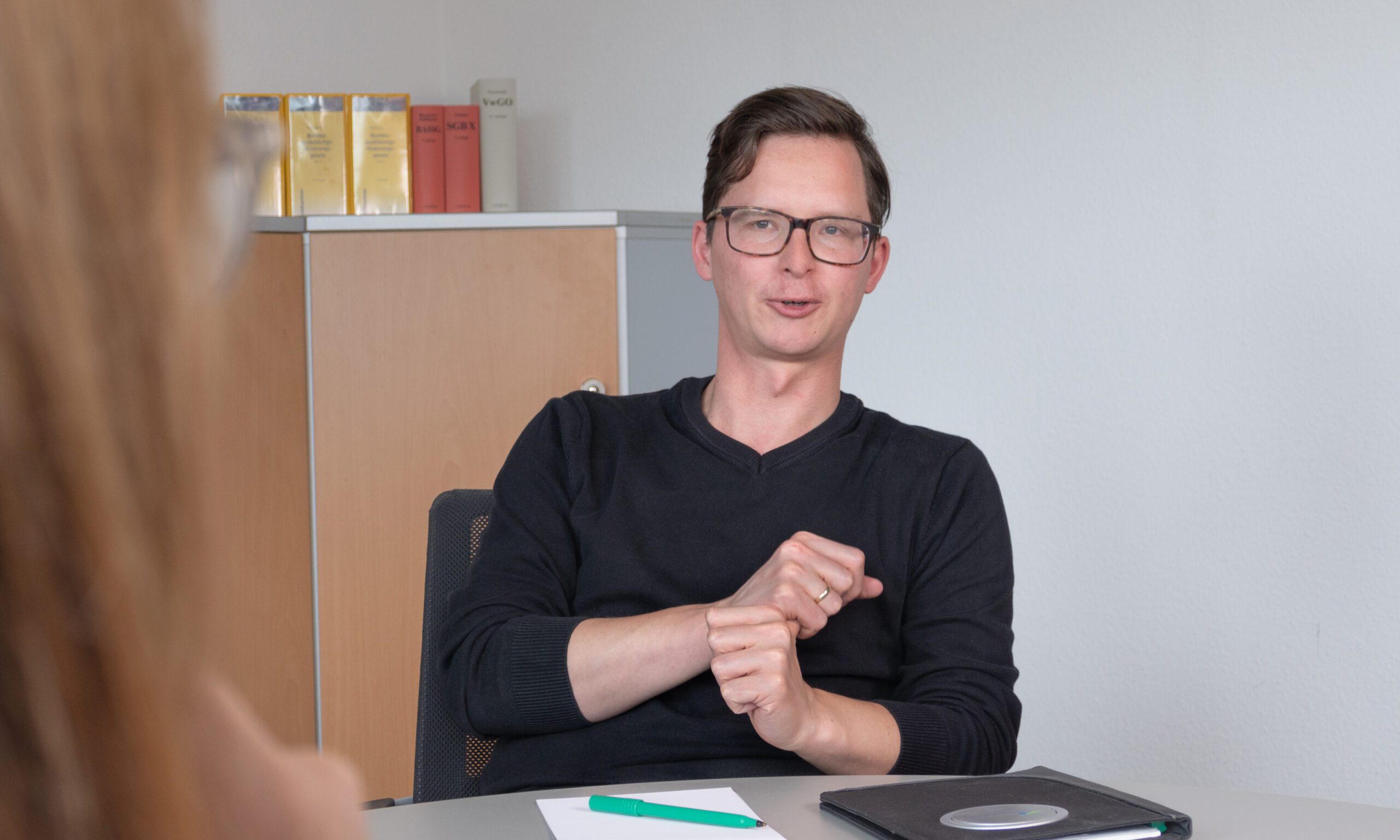 Ein Mann mit Brille und schwarzem Pullover sitzt an einem Schreibtisch und gestikuliert während eines Gesprächs mit den Händen. Auf dem Tisch liegen ein Notizblock mit einem grünen Stift und ein geschlossener Laptop. Eine verschwommene Person steht ihm gegenüber.