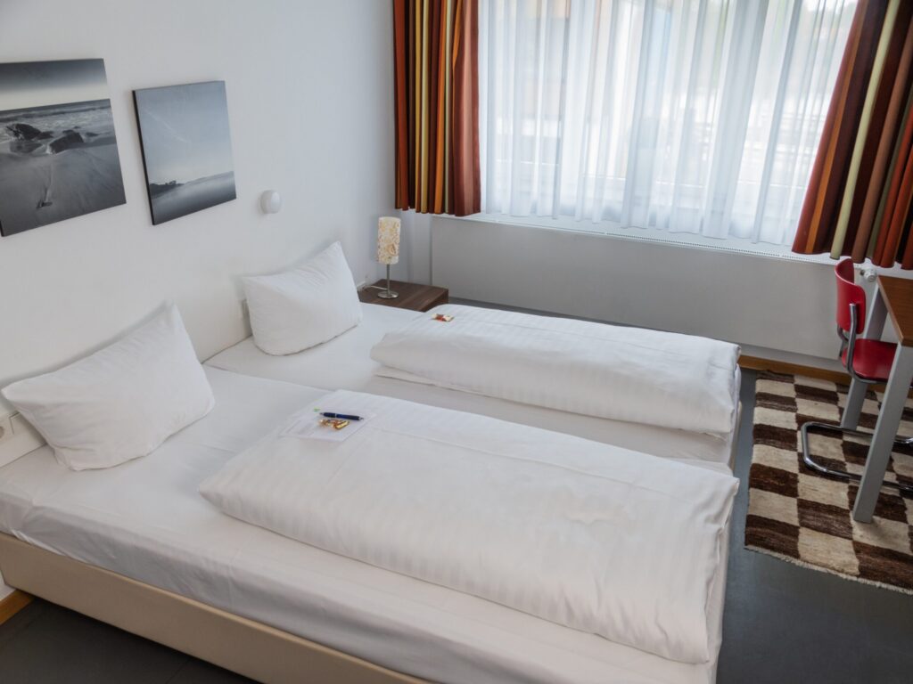 A hotel room with two clean single beds with white bed linen, a bedside table with a lamp between them, a window with striped curtains, wall art, a brown carpet and a red chair at a desk.