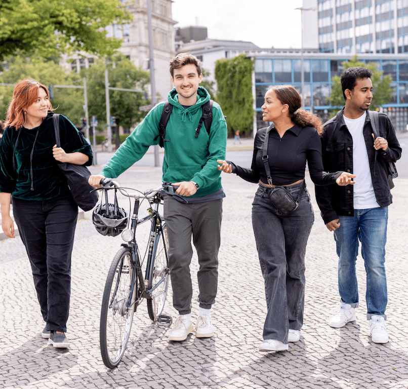 Vier junge Erwachsene gehen zusammen im Freien spazieren und unterhalten sich; eine Person schiebt ein Fahrrad. Sie scheinen glücklich zu sein und sich zu unterhalten, während im Hintergrund Gebäude und Bäume zu sehen sind.